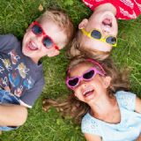 Tre små grinende børn med solbriller på ligger på en græsplæne.